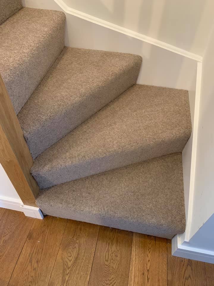 Carpeted stairs with sharp triangular steps leading up from a wooden floor, turning at a right angle against white walls and a wooden banister.