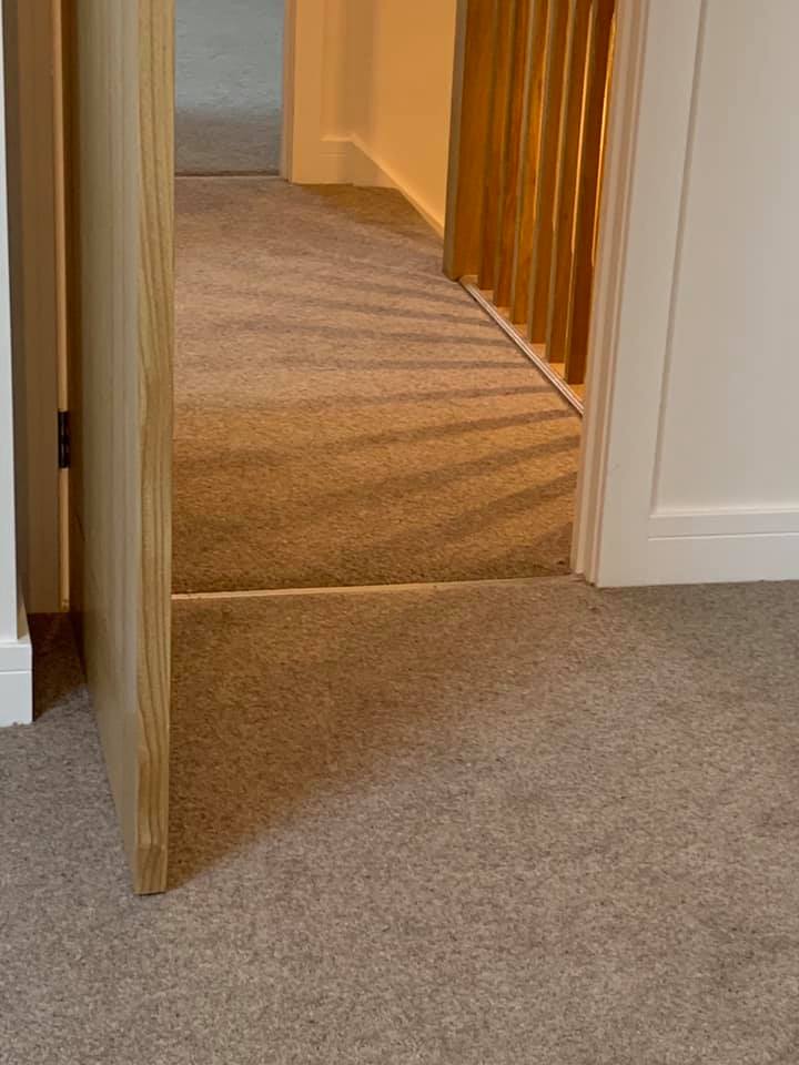 An open wooden door leads to a hallway with beige carpet. Sunlight through wooden railing casts striped shadows on the carpet near the door and wall.