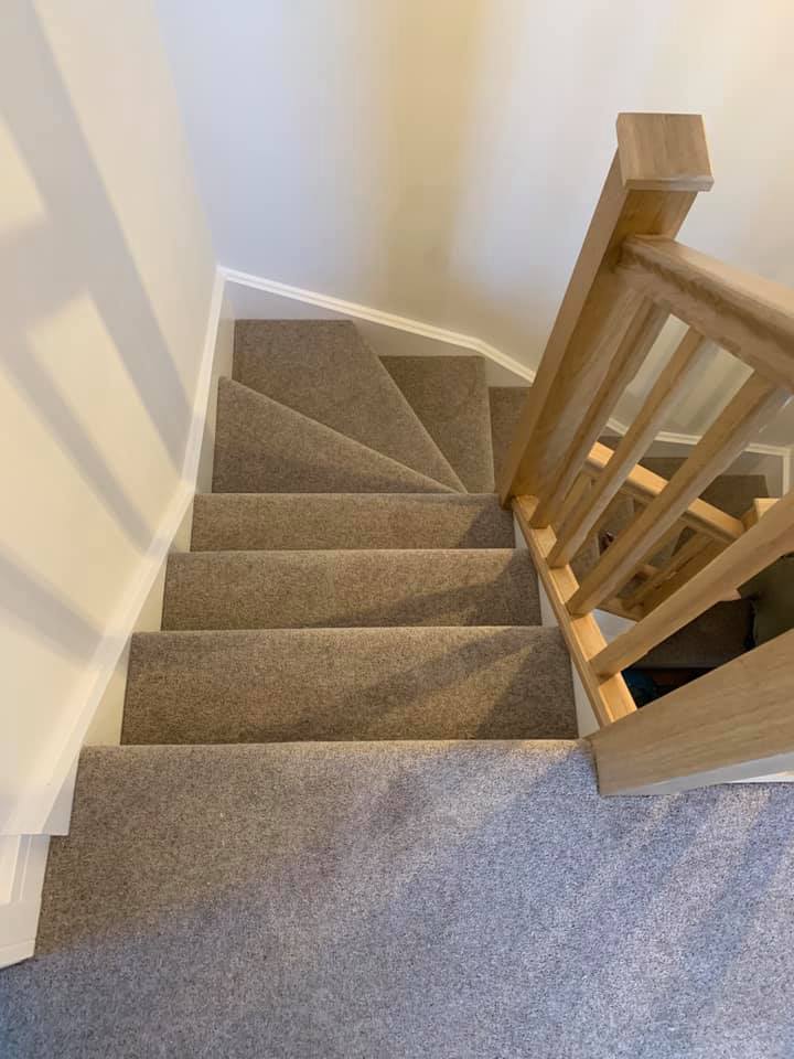 A carpeted staircase with a turn, featuring wooden railings on the right and cream-colored walls on the left. The stairs are seen from the top, looking downward.