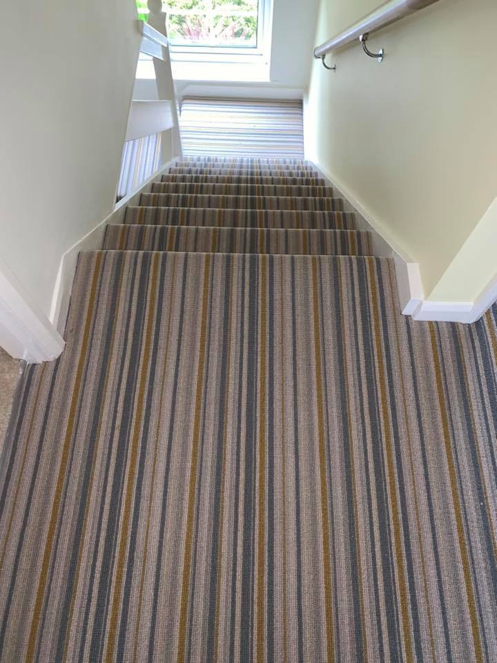 View looking down a staircase covered with a striped carpet in shades of beige, gray, and brown; a handrail is mounted on the right wall and a window with natural light is visible at the bottom.