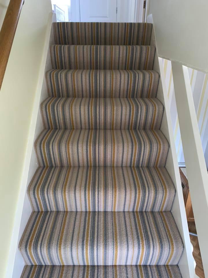 A staircase covered with a striped carpet in beige, blue, green, and mustard yellow tones, creating an optical illusion that makes the steps appear uneven or distorted. White railings border the stairs.