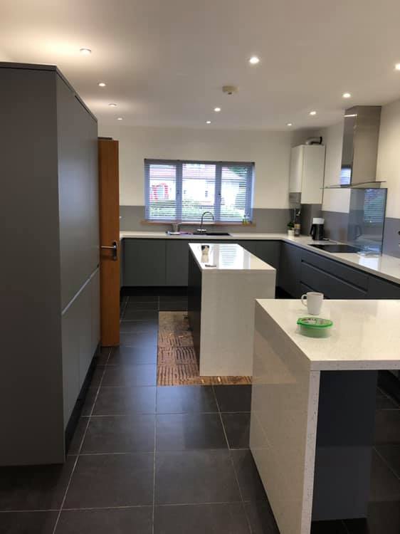 Modern kitchen with dark cabinets, white countertops, an island, and built-in appliances. A window above the sink lets in natural light. There is a green ashtray and a mug on the island. The floor is dark tile.