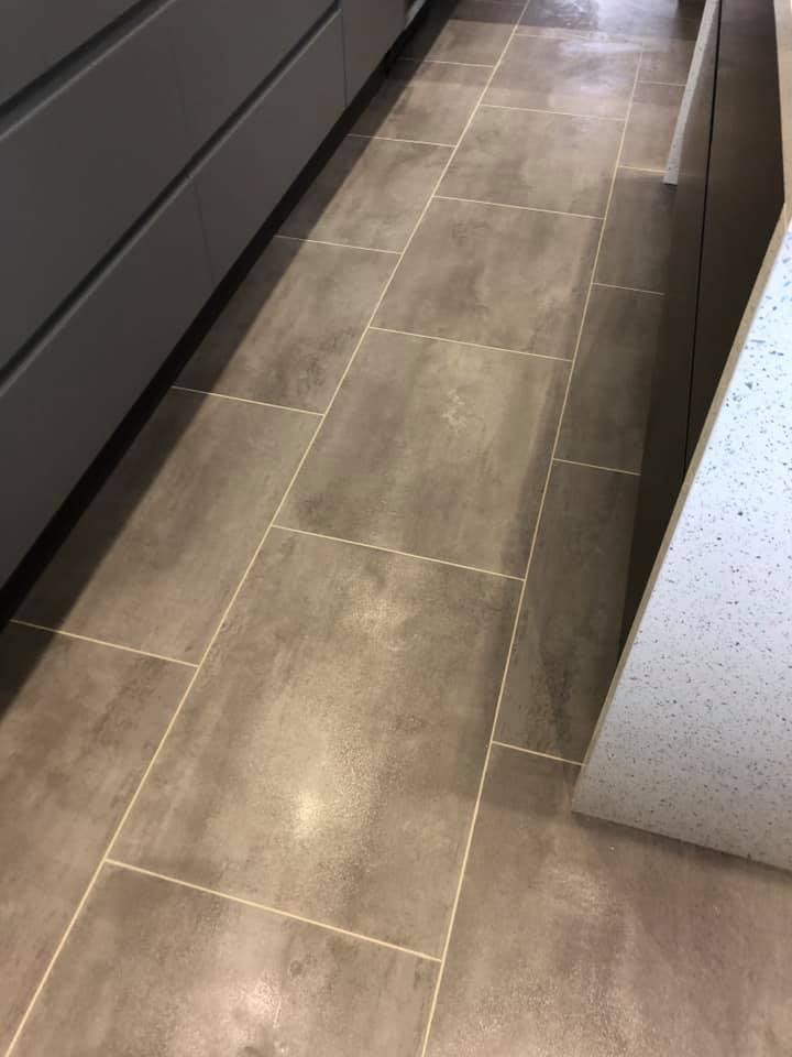 A kitchen floor with large rectangular gray tiles arranged in a staggered pattern. Dark cabinets are on the left and a light speckled countertop is on the right.