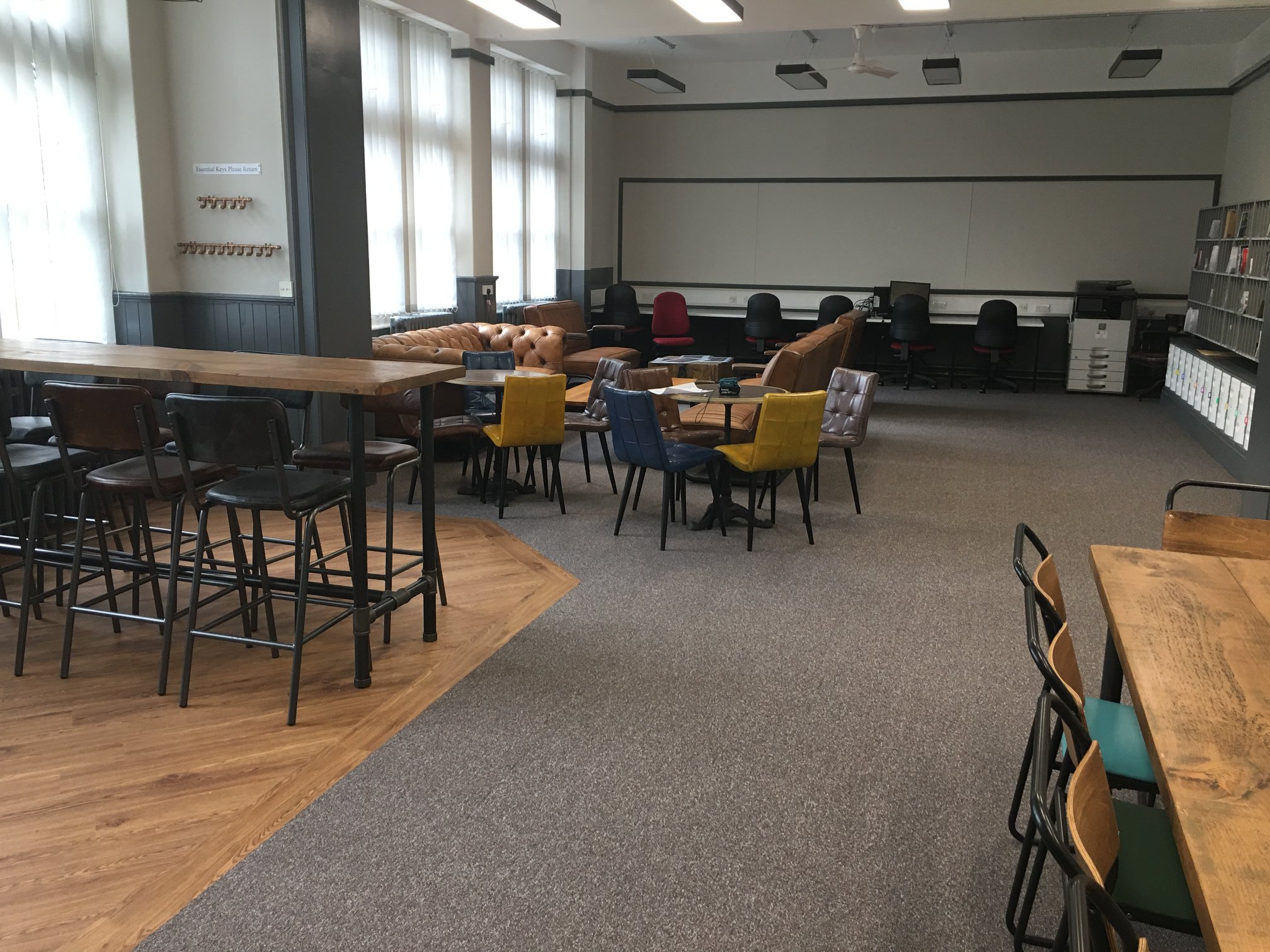 A spacious, modern room with tall windows, various seating areas including sofas, armchairs, and high tables with stools, bookshelves on one wall, and computer stations along the back.