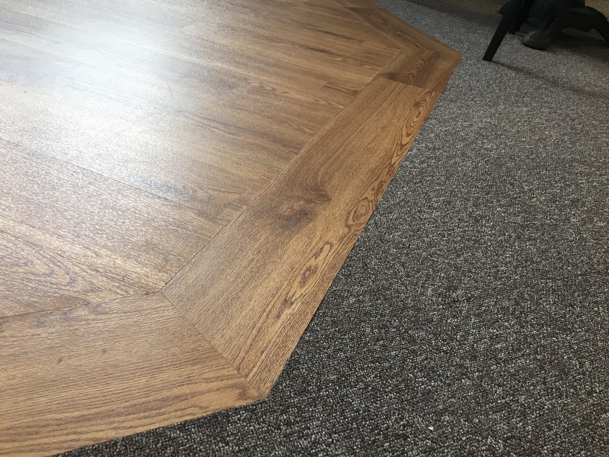 Close-up of a corner where light brown wooden flooring meets textured grey carpet, separated by a diagonal edge of the wood. The leg of a chair and part of a black bag are visible in the background.