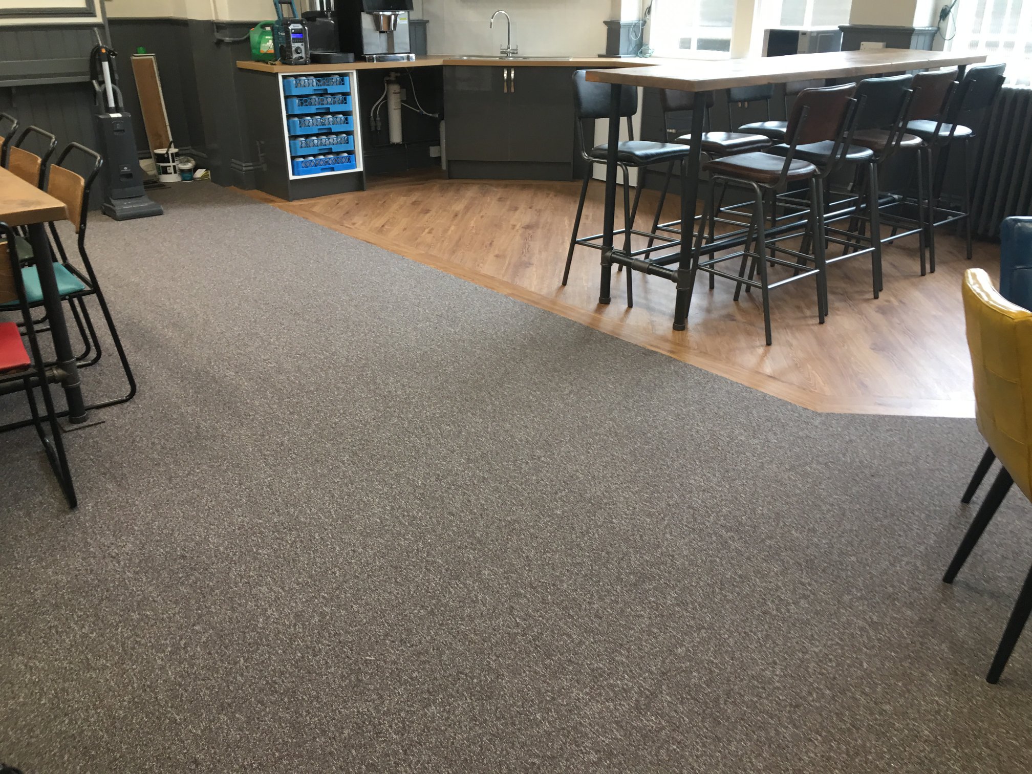 Office kitchen area with gray carpet meeting wood flooring, high tables and chairs, a coffee station, sink, blue storage shelf, and various chairs around. Bright and modern atmosphere with natural light from windows.