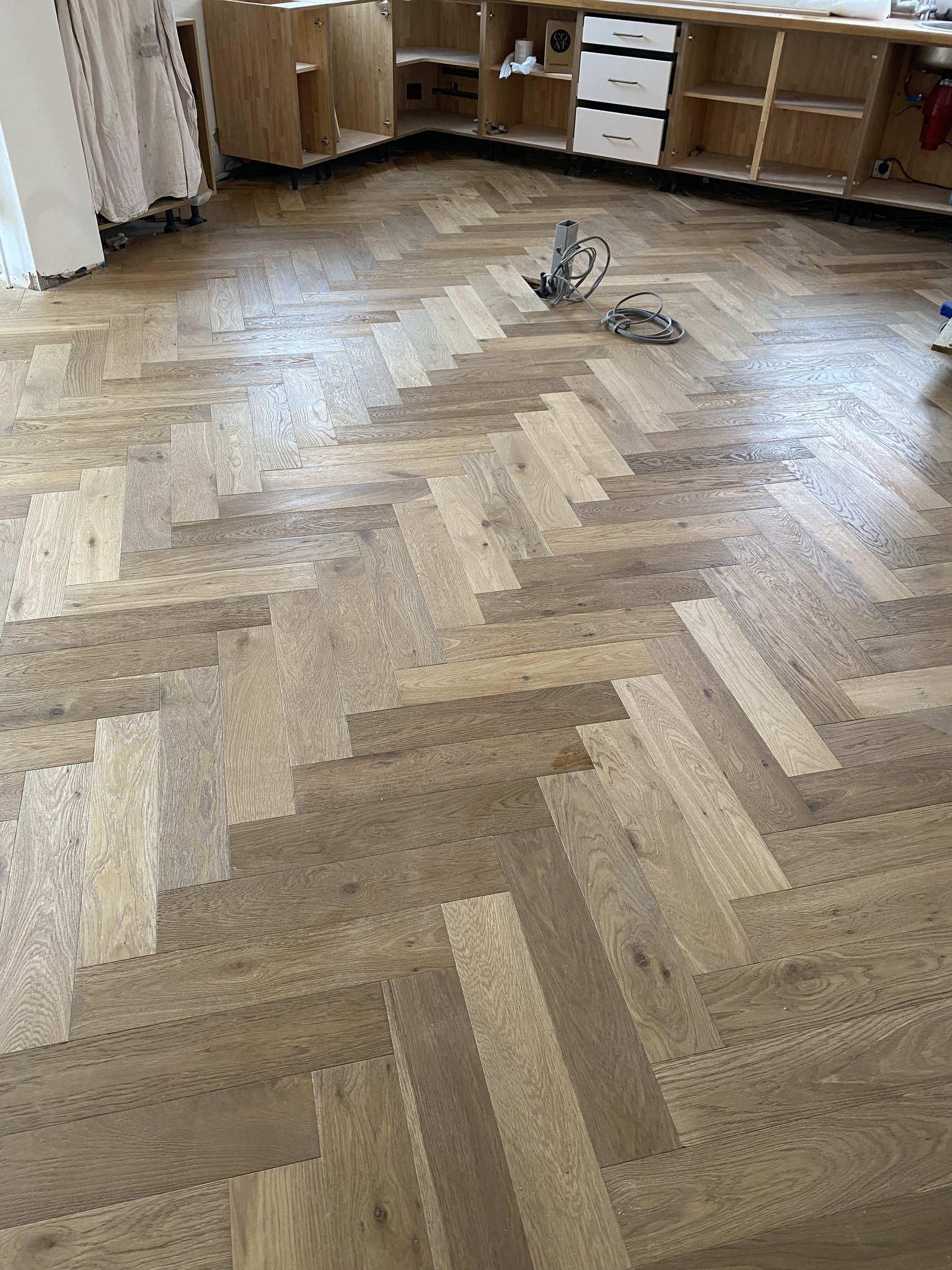 A room with newly installed herringbone-patterned wooden flooring. Some tools and cables lie on the floor, and unfinished wooden kitchen cabinets are visible in the background.