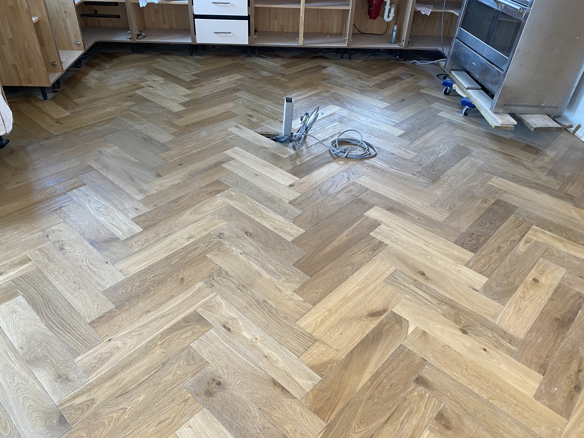 A kitchen under renovation with unfinished cabinets and a newly installed herringbone-patterned wooden floor. Loose wires and a pipe protrude from the center of the floor.