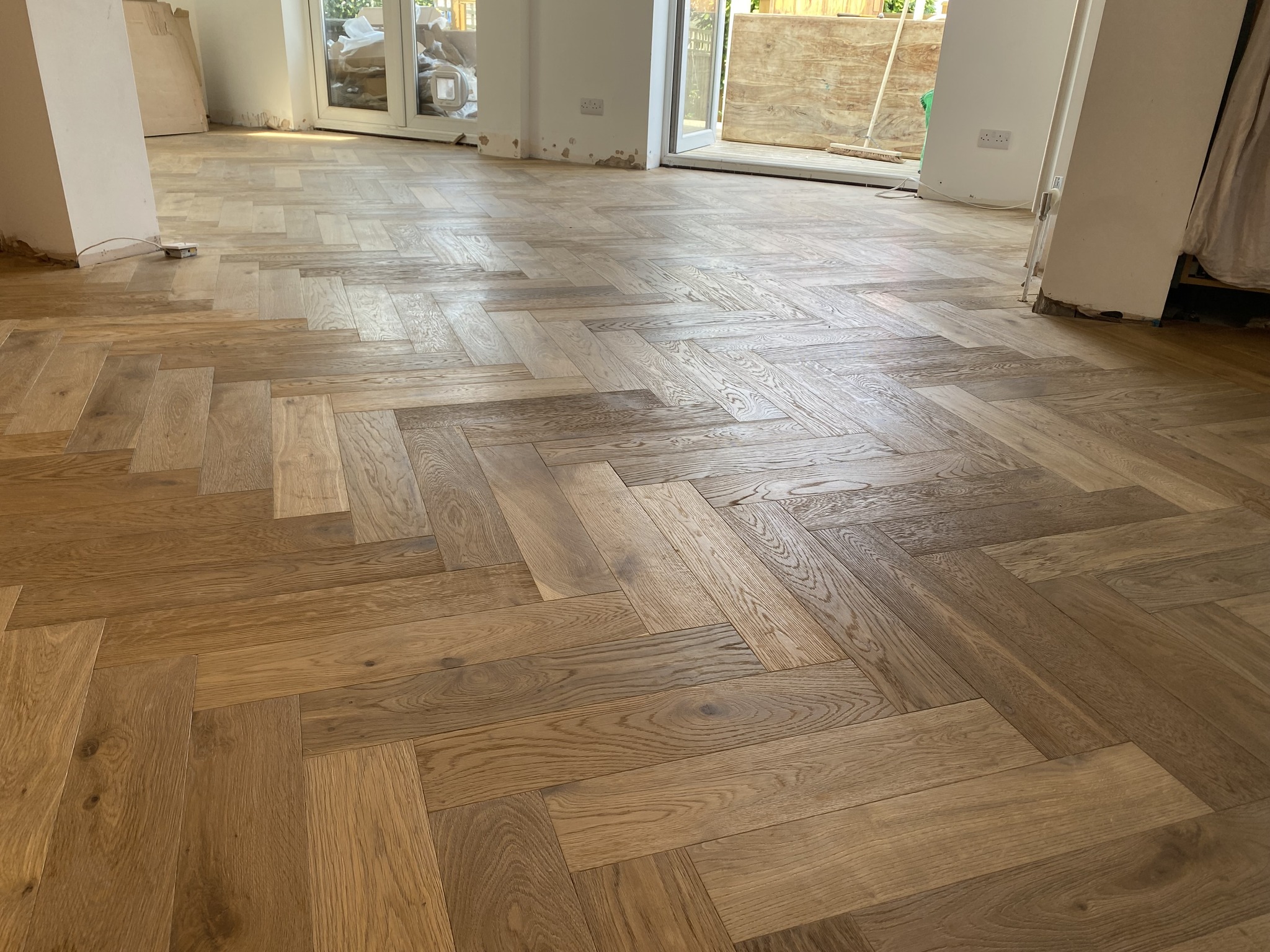 Herringbone patterned wooden flooring in a bright, unfurnished room with large glass doors and some construction materials visible in the background.