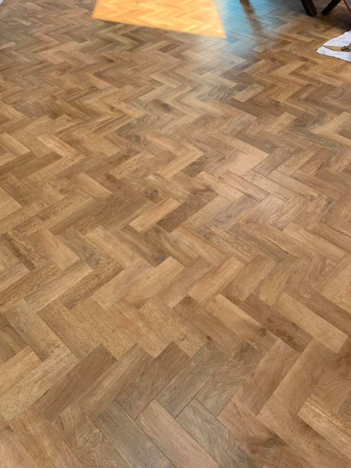 A sunlit wooden floor with a herringbone pattern. Warm light highlights the parquet design, with shadows and sunlight visible near the top corner.