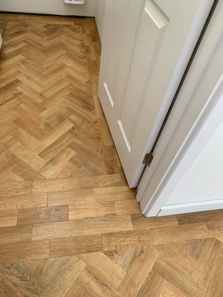 A room corner with light wooden flooring in a herringbone pattern abruptly meeting straight-laid planks, next to a white door and white walls.
