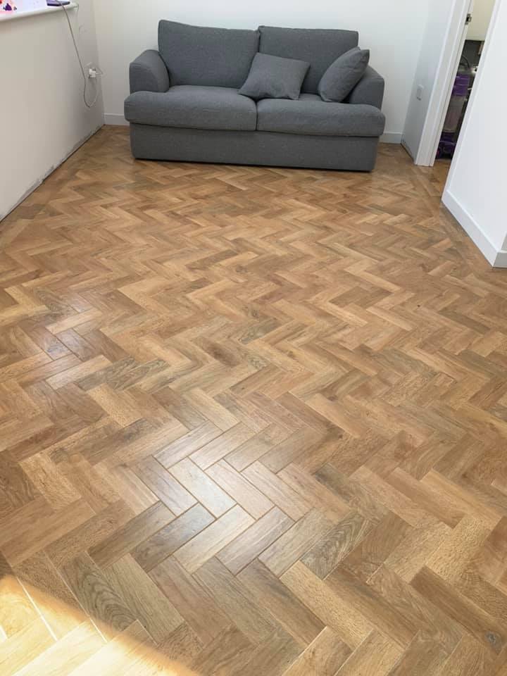 A small room with wooden herringbone-patterned flooring, featuring a grey sofa with a cushion and two pillows against a white wall.