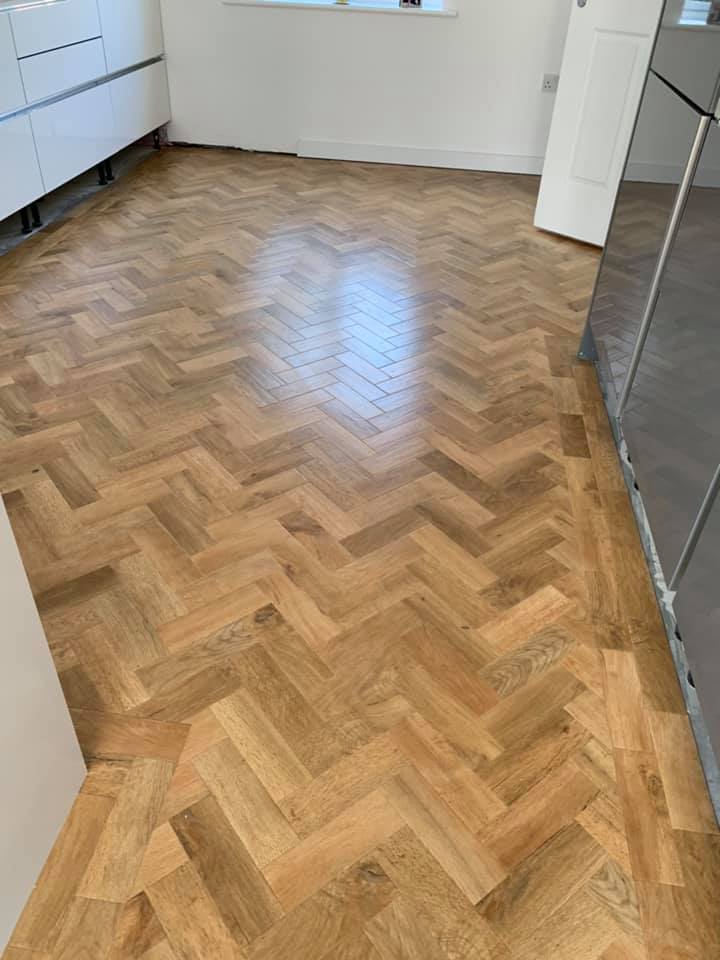 A kitchen with light wood herringbone patterned flooring, white cabinets on the left, and a window at the far end letting in natural light.