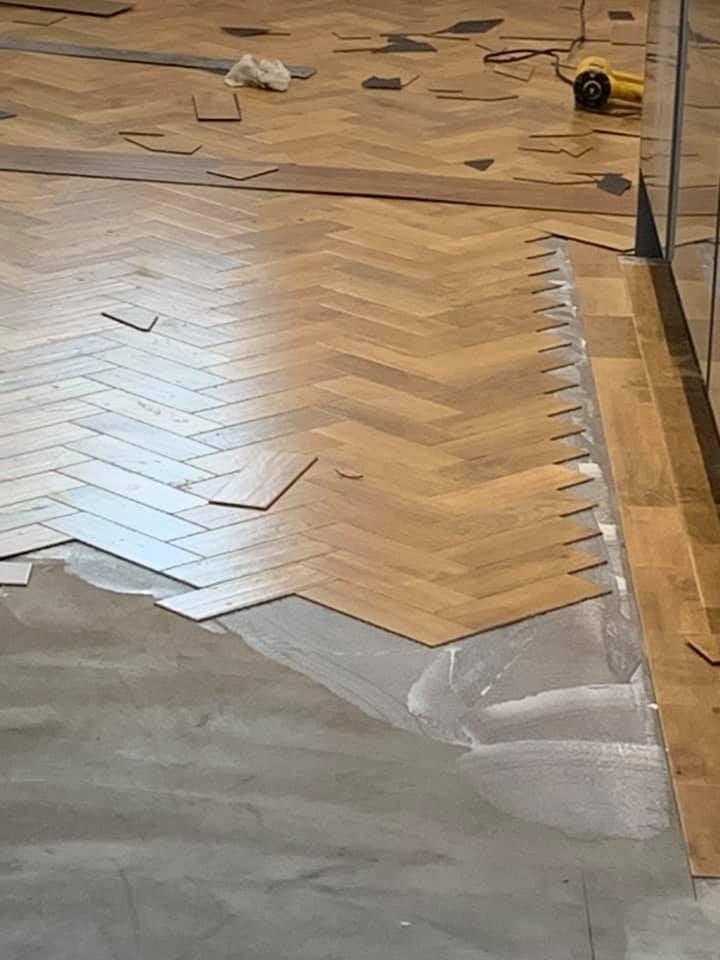 Partially installed herringbone-patterned wooden flooring, with adhesive visible on the unfinished section; tools and tile pieces scattered nearby. A glass wall runs along the right side of the image.