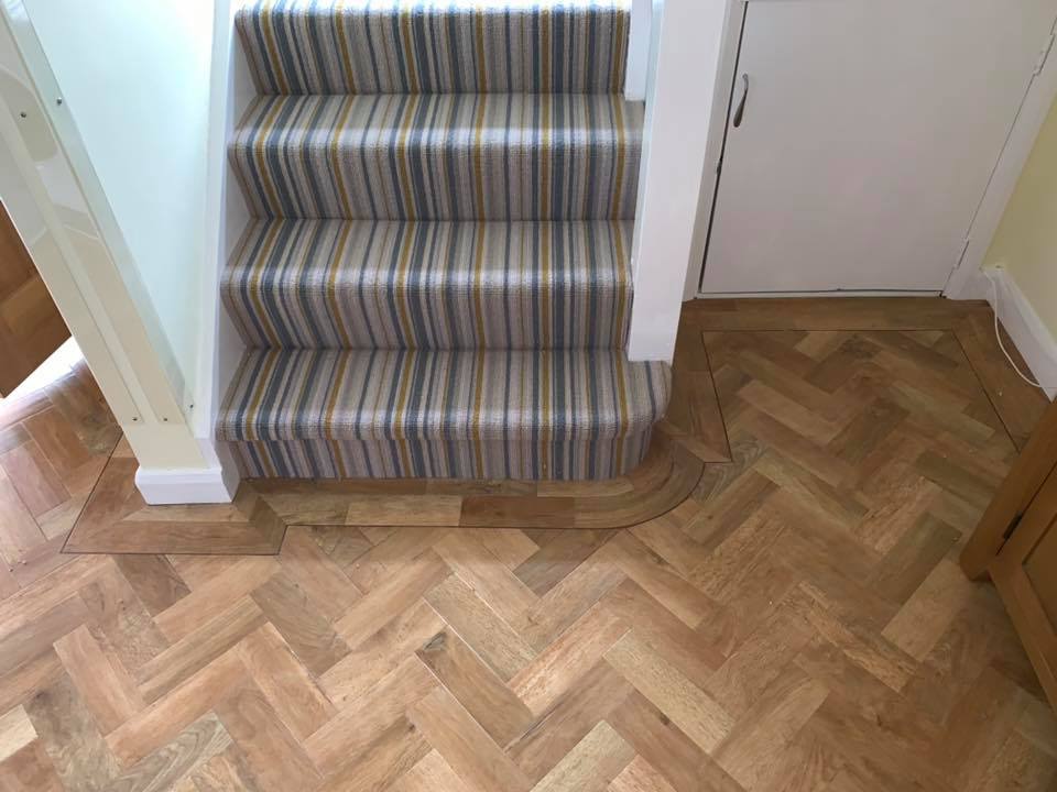 A staircase with striped carpet leads down to a hallway with herringbone-patterned wood flooring featuring a curved edge where it meets the stairs. A white door is visible near the stairs.