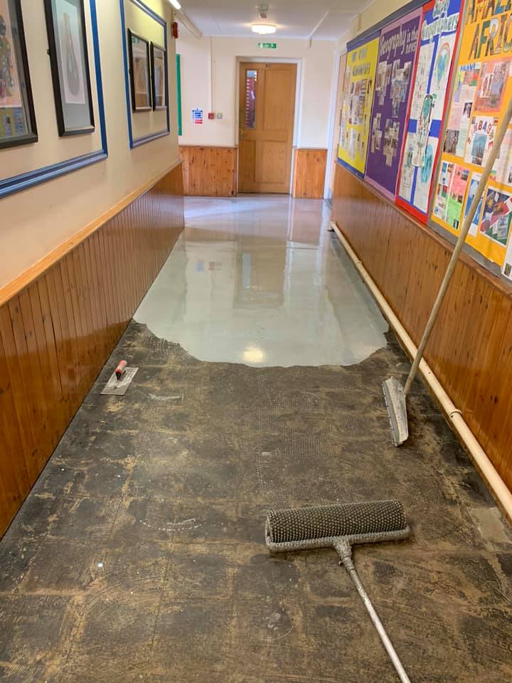 A hallway with colorful posters on the walls is being resurfaced; the floor is partly covered in fresh, wet concrete, with resurfacing tools lying on the unfinished section.
