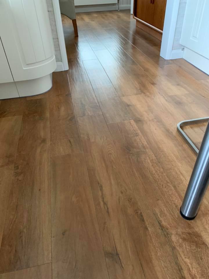 Wooden laminate flooring in a kitchen or dining area, with white cabinets, a metal chair leg, and part of a beige upholstered chair visible. The floor has a natural brown wood grain pattern.