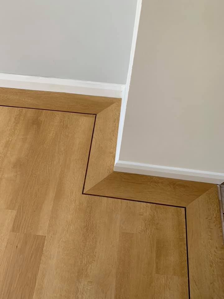 Light wood flooring with a border that awkwardly follows the shape of a protruding corner in the white wall, creating an unusual and irregular angled pattern around the corner.