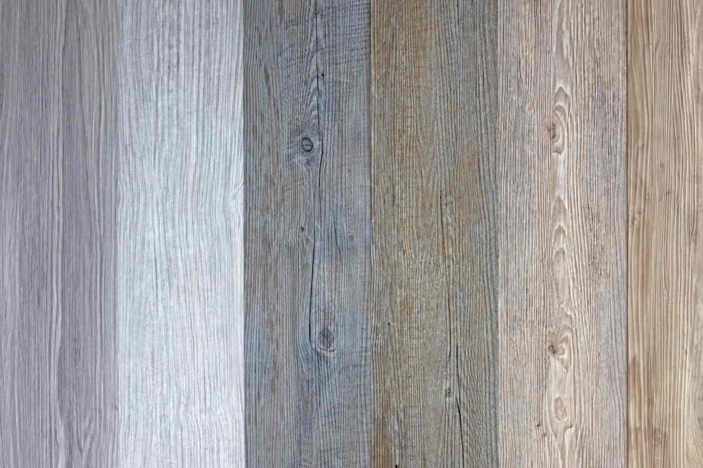 Vertical wooden planks lined up side by side, each with a different shade ranging from light gray to medium brown, displaying various wood grain patterns and textures.