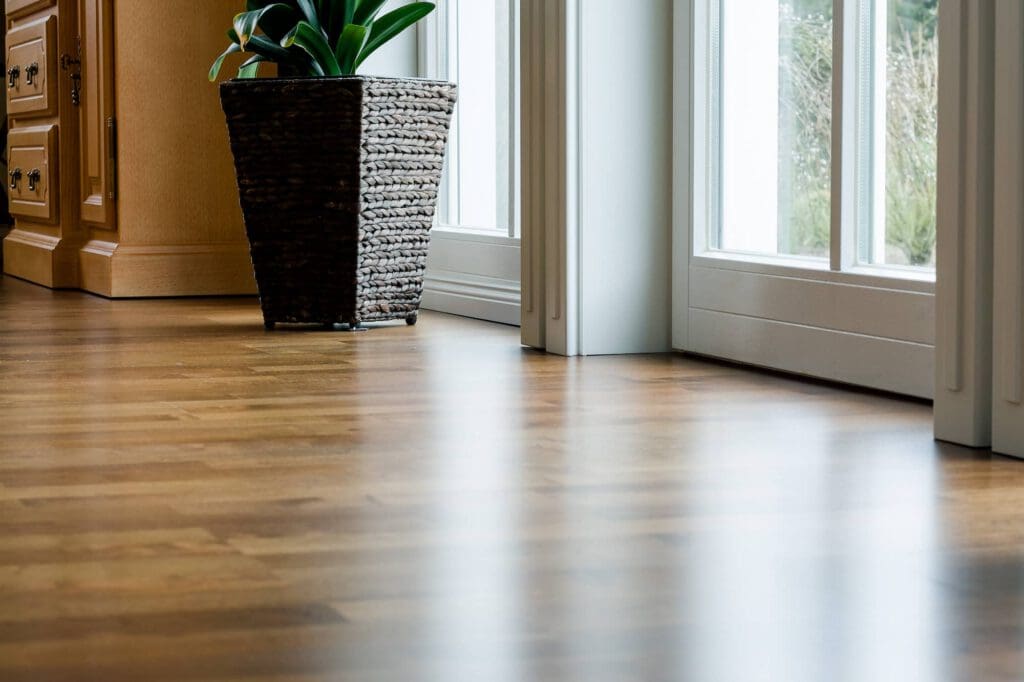 A close-up view of a clean wooden floor with sunlight streaming in through large glass doors. A woven basket planter with a green plant sits near the doors, next to a wooden cabinet.