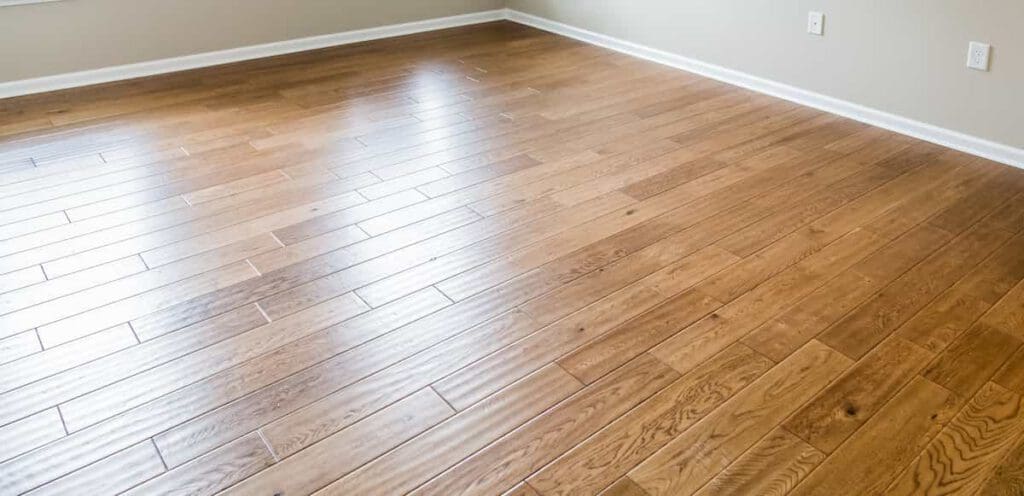 Empty room with light brown hardwood flooring, beige walls, white baseboards, and two electrical outlets visible on the right wall. Sunlight is reflecting off the floor near the windows.